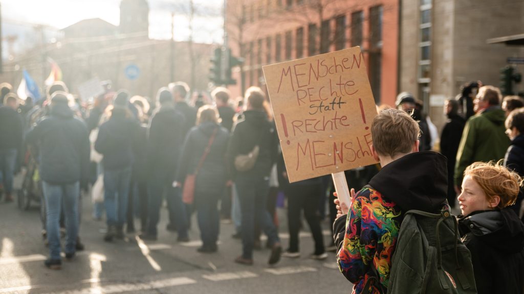 Menschen auf Demonstration mit einem Plakat im Fokus, welches den Titel Menschenrechte statt rechte Menschen trägt